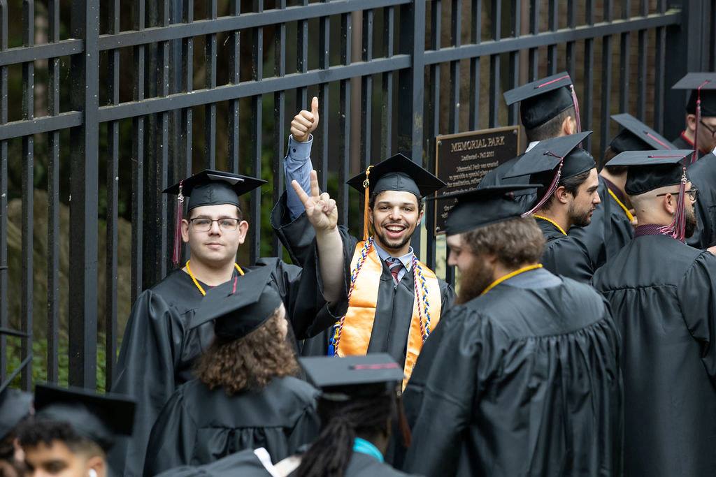 Two students smile at the camera during a graduation procession. One gives a thumbs up. 